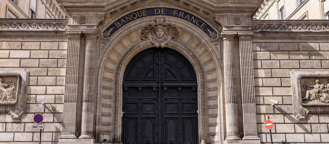 Vor dem Eingang der Banque de France in Paris steht ein Mann an einer Mauer. | AFP Vor dem Eingang der Banque de France in Paris steht ein Mann an einer Mauer.