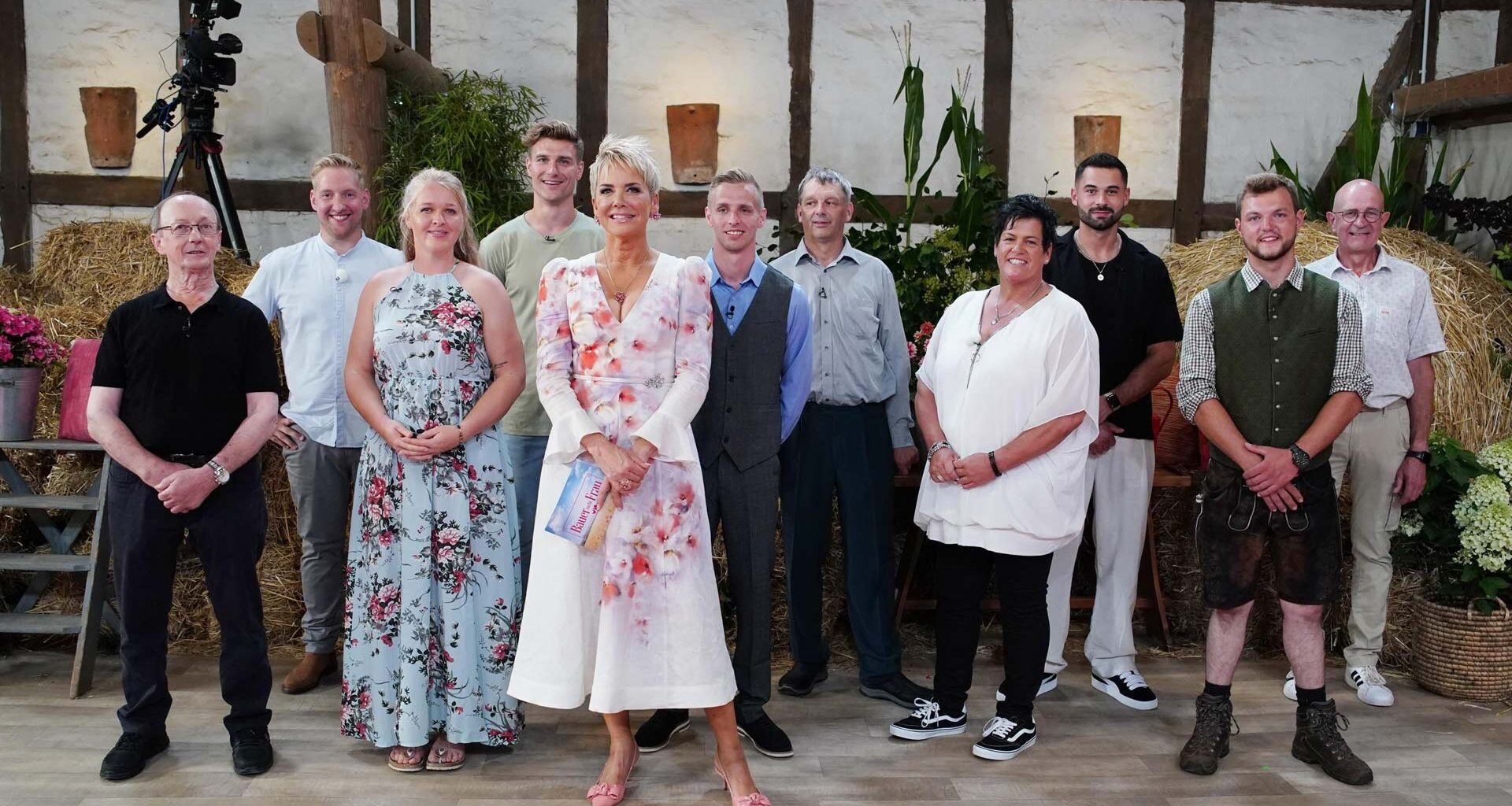 Auf dem Bild zur Sendung Bauer sucht Frau 2025 stehen die Moderatorin Inka Bause und die Teilnehmer:innen Walter, Daniel, Insa, Friedrich, Christopher, Johann, Simone, Basti, Michi und Herbert in einem rustikal dekorierten Studio mit Strohballen und Blumen.