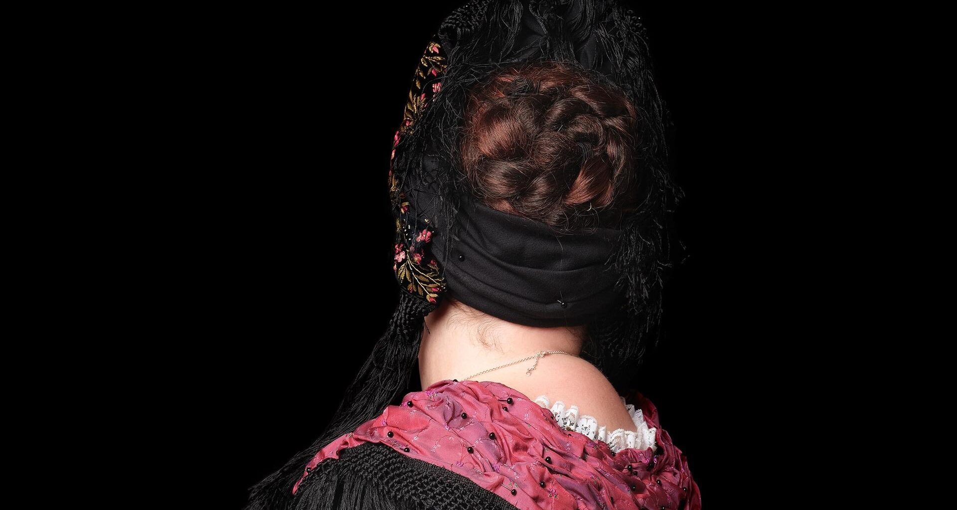 Rückenansicht einer Frau in Trachtenkleidung mit einem weißen Rock, schwarzer Bluse und verziertem rotem Schal.