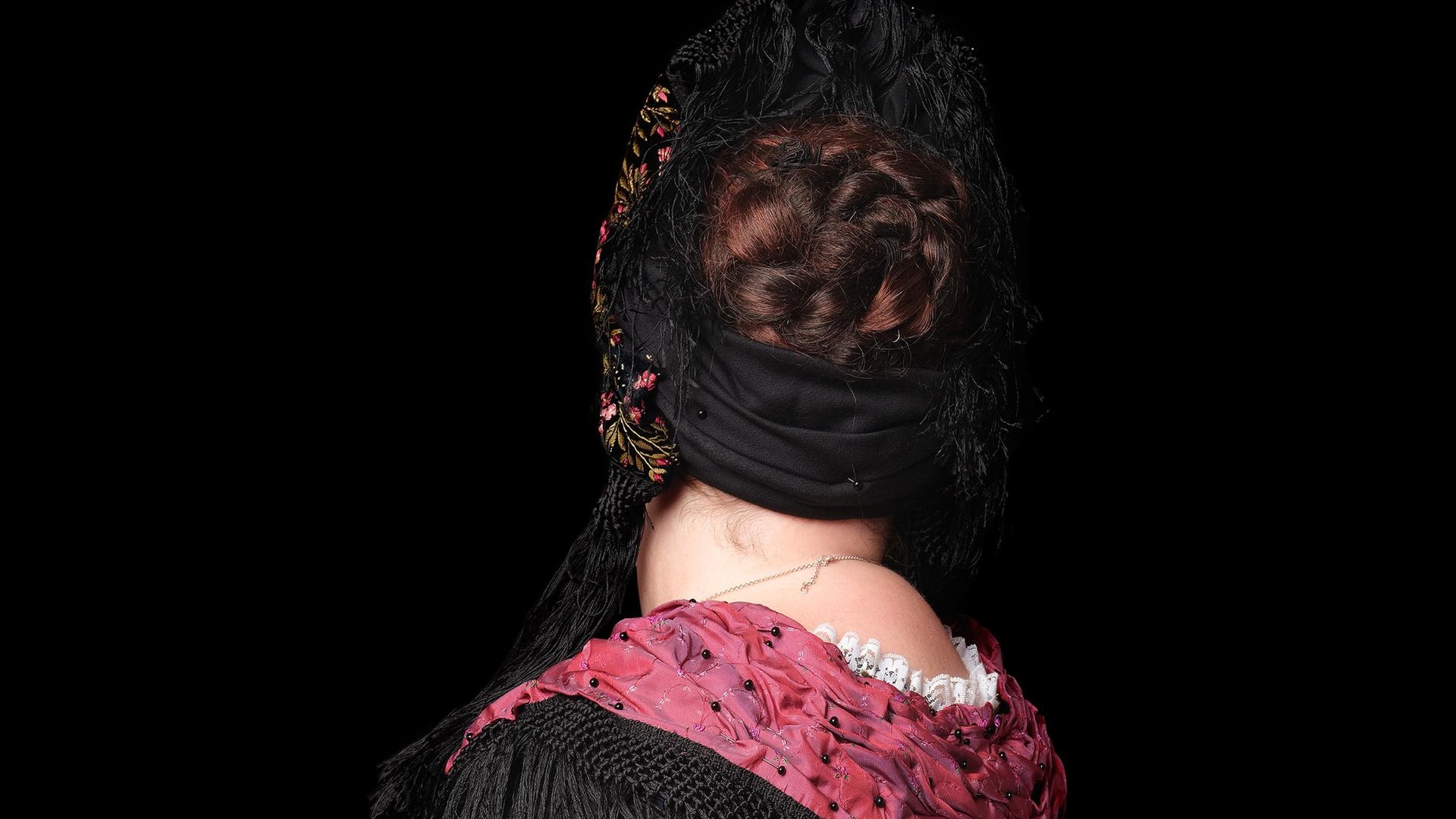 Rückenansicht einer Frau in Trachtenkleidung mit einem weißen Rock, schwarzer Bluse und verziertem rotem Schal. 