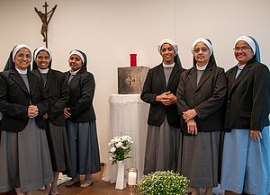 Sechs Ordensschwestern stehen nebeneinander in einem schlicht eingerichteten Raum mit einem Kruzifix an der Wand. Alle tragen graue Kleider mit schwarzem Blazer und Schleier. In der Mitte des Raumes steht ein kleiner Altar mit einem Tabernakel, darüber eine rote Lampe. Auf dem Boden stehen weiße Blumen und eine Kerze.