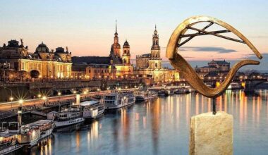 ie beleuchtete Silhouette der Dresdner Altstadt bei Sonnenuntergang, einschließlich der Frauenkirche und der Hofkirche, gespiegelt in der Elbe. Im Vordergrund rechts steht eine moderne, bronzefarbene Skulptur auf einem Steinsockel.