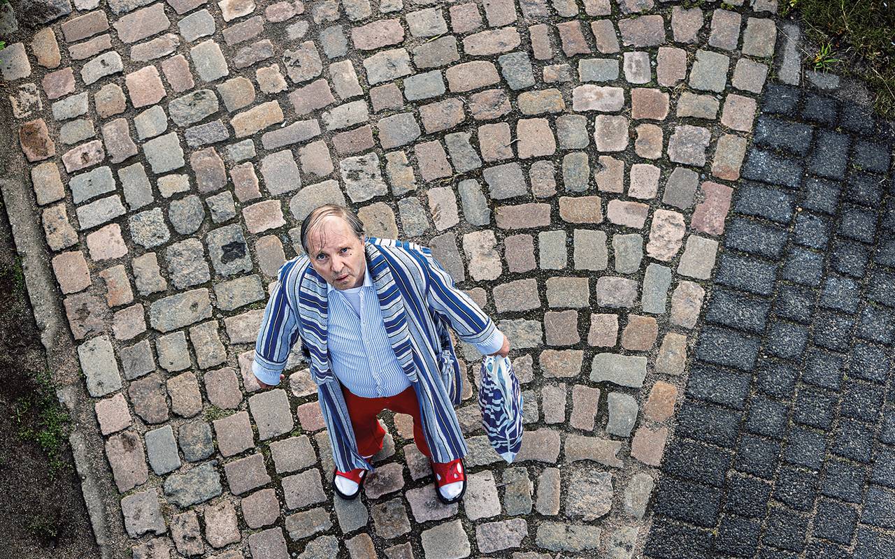 "Dittsche" steht im Bademantel, Latschen und mit Aldi-Tüte in der Hand auf einem Kopfsteinpflaster.