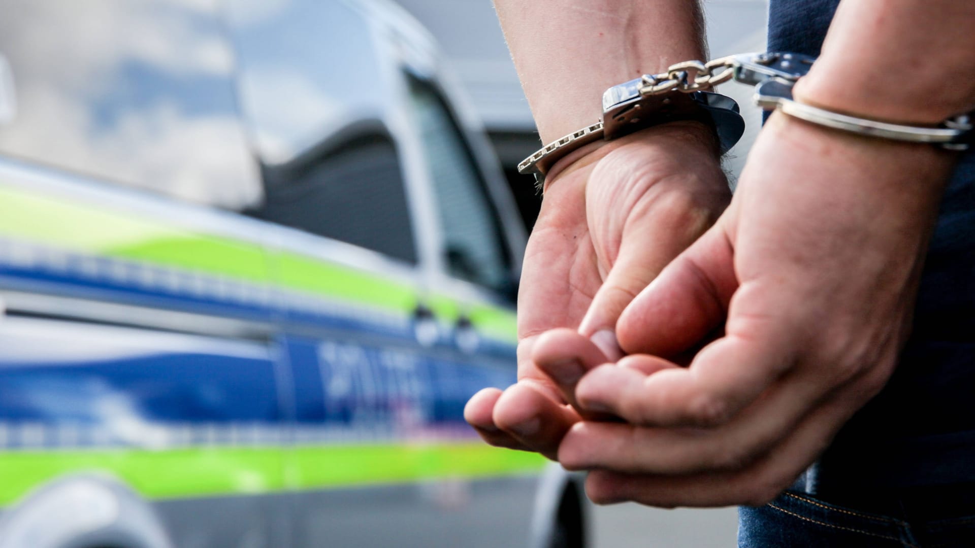 Ein Mann steht mit Handschellen gefesselt neben einem Streifenwagen (Archivbild): Fünf der festgenommenen Personen sitzen jetzt in Untersuchungshaft.