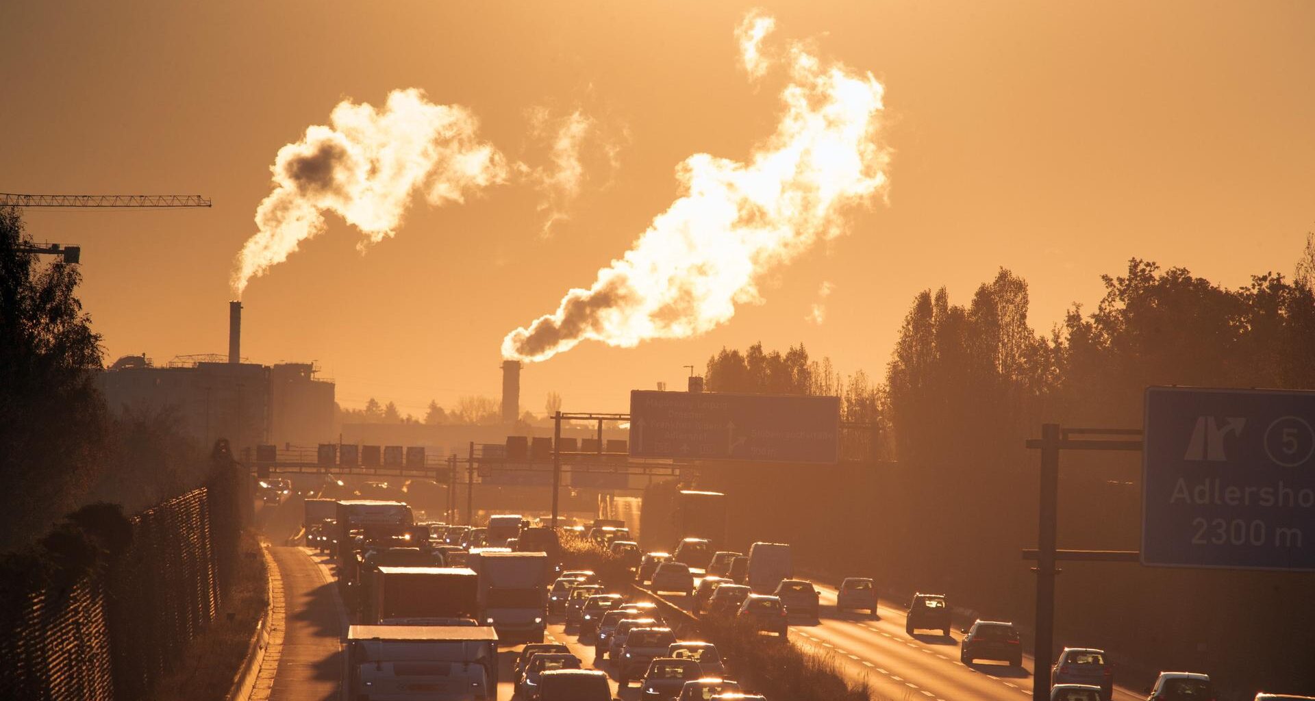 Stau am Morgen auf der Autobahn, im Gegenlicht sind im Hintergrund Rauschschwaden aus Industrieschornsteinen zu erkennen.