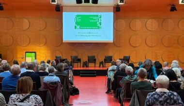 Publikum in einem Saal, Bühne mit vier leeren Stühlen, Leinwand mit Begrüßungstext.