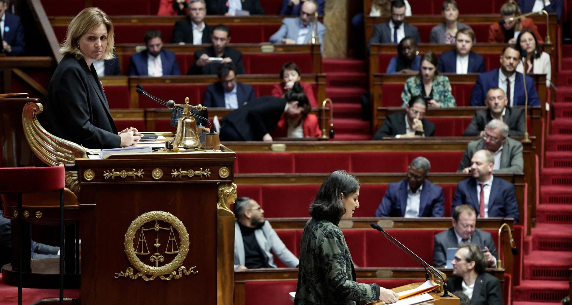 Man sieht im Vordergrund eine Frau im erhöhten Parlalementspräsidium und eine weitere Frau am Rednerpult jeweils von der Seite. Dahinter unscharf Abgeordnete auf den roten Bänken.