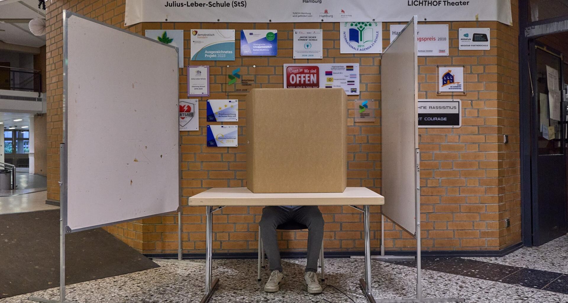 Blick auf eine Wahlkabine in einer Schule in Hamburg. Ein Mann sitzt an einem Tisch. Er wird von einem Sichtschutz verdeckt.