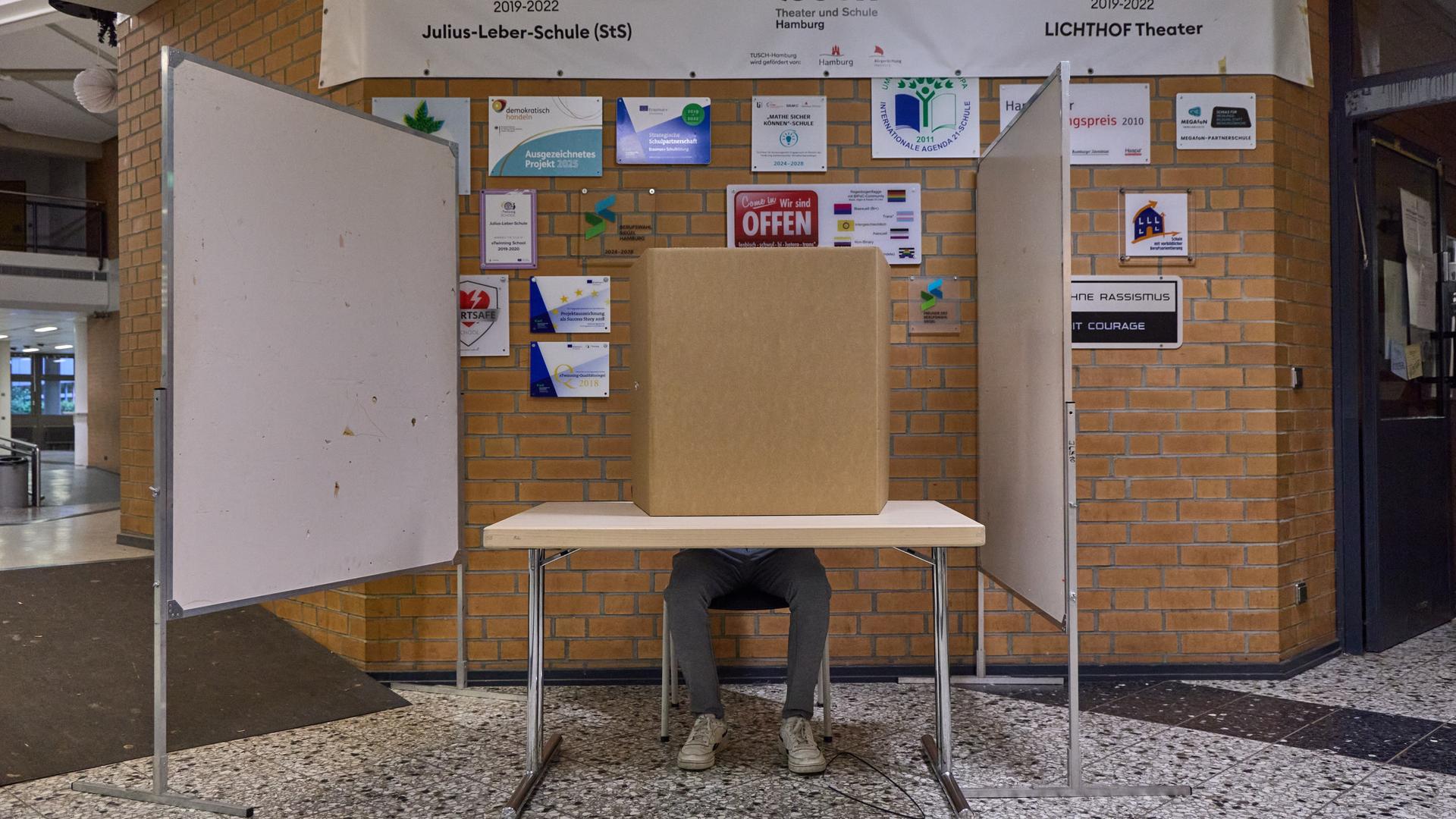 Blick auf eine Wahlkabine in einer Schule in Hamburg. Ein Mann sitzt an einem Tisch. Er wird von einem Sichtschutz verdeckt. 