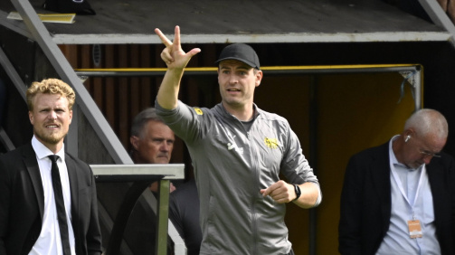 © imago - Das Foto zeigt die Trainerbank eines Fußballstadions während eines Spiels. Im Vordergrund stehen zwei Männer. Links steht ein Mann in einem schwarzen Anzug mit weißem Hemd und schwarzer Krawatte: Trainer Oscar Hiljemark von Elfsborg. Er hat lockige, blonde Haare und blickt konzentriert nach vorne. Rechts daneben steht ein Mann in Sportkleidung von Mjällby AIF, Trainer Karl Marius Aksum – graues Oberteil mit Vereinslogo, schwarze Trainingshose und schwarze Kappe. Er hebt den rechten Arm und zeigt mit der Hand eine Geste, die an eine taktische Anweisung erinnert. Im Hintergrund sind weitere Personen zu sehen, darunter ein älterer Mann mit grauen Haaren und Brille, der auf sein Handy schaut. Die Szene spielt offenbar während eines laufenden Fußballspiels, und die Stimmung wirkt konzentriert und angespannt.