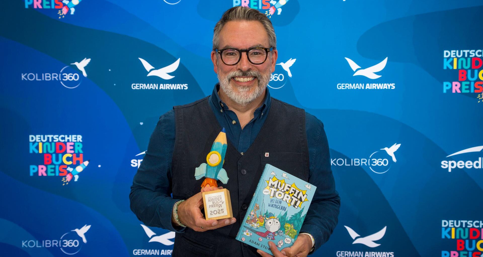 Der Autor Adam Stower hält den deutschen Kinderbuchpreis in einer Hand, in der anderen sein Buch "Muffin und Tört".