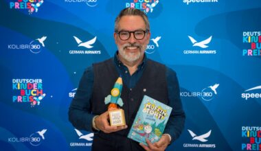 Der Autor Adam Stower hält den deutschen Kinderbuchpreis in einer Hand, in der anderen sein Buch "Muffin und Tört".