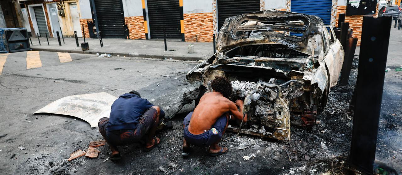 Menschen ducken sich hinter einem ausgebrannten Polizeiauto in Rio de Janeiro. | EPA Menschen ducken sich hinter einem ausgebrannten Polizeiauto in Rio de Janeiro.