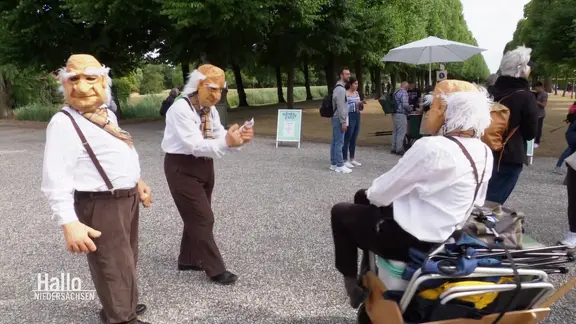 Drei Personen in Kostümen und mit großen Pappmaché-Köpfen sowie Passanten in einem Park.