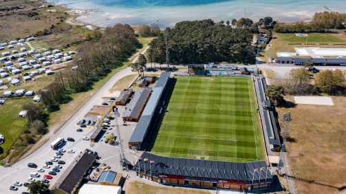 © imago - Das Bild zeigt eine Luftaufnahme des Fußballstadions Strandvallen, der Heimspielstätte des schwedischen Vereins Mjällby AIF. Das Stadion liegt direkt an der Ostseeküste, mit dem Meer und einem hellen Sandstrand im Hintergrund. Der grüne Rasen des Spielfelds ist gut gepflegt und rechteckig von Tribünen umgeben. Die Haupttribüne befindet sich im unteren Bildbereich und ist mit einem schwarzen Dach gedeckt. Auf dem Dach sind Flaggen und Werbeschilder zu sehen. Neben dem Stadion liegen Parkplätze mit mehreren Autos, einige Gebäude sowie ein Campingplatz mit vielen weißen Wohnwagen. Im Hintergrund erstrecken sich Wälder und die ruhige, blaue Ostsee. Die Atmosphäre wirkt ruhig und sonnig.