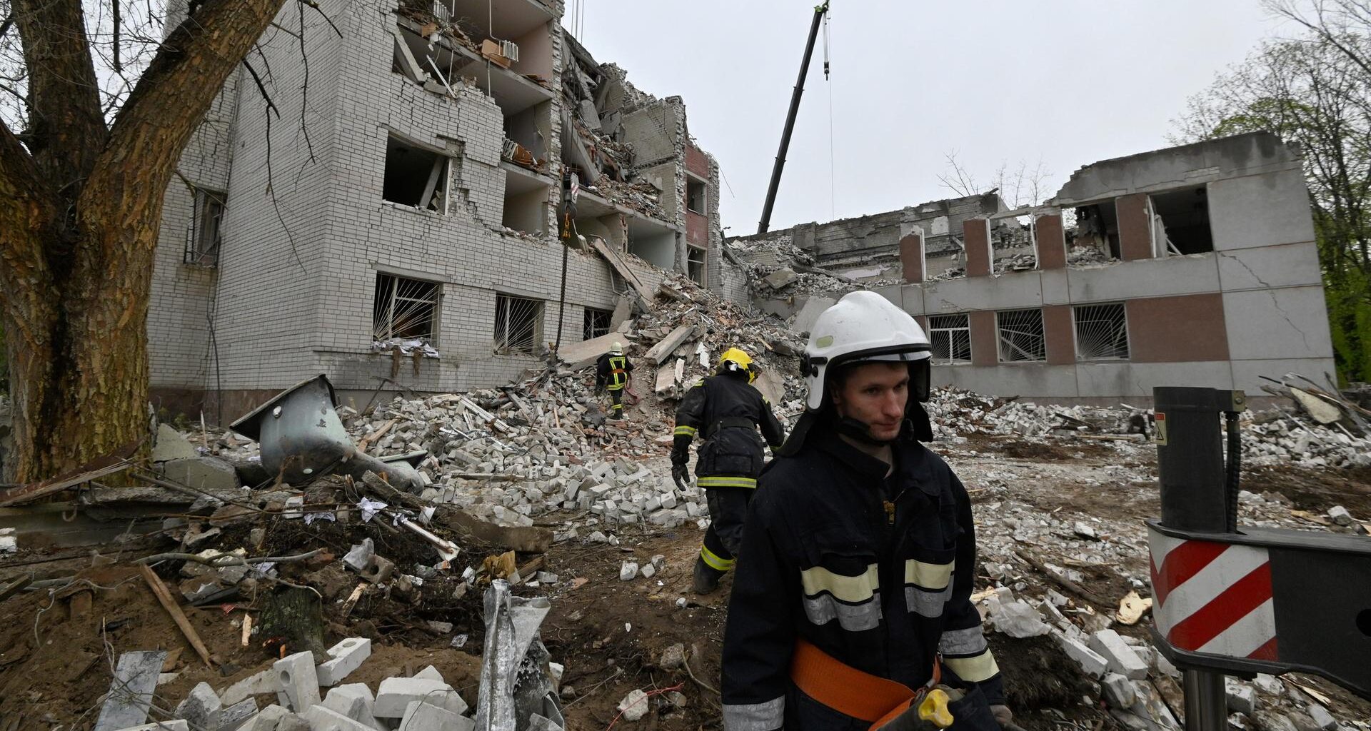 Zwei Feuerwehrleute arbeiten vor den Trümmern eines eingestürzten Hauses. Der Himmel ist bedeckt.