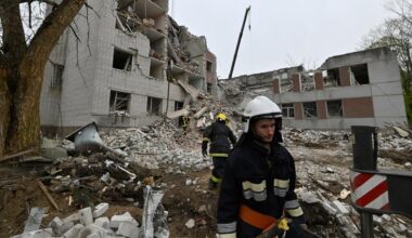 Zwei Feuerwehrleute arbeiten vor den Trümmern eines eingestürzten Hauses. Der Himmel ist bedeckt.