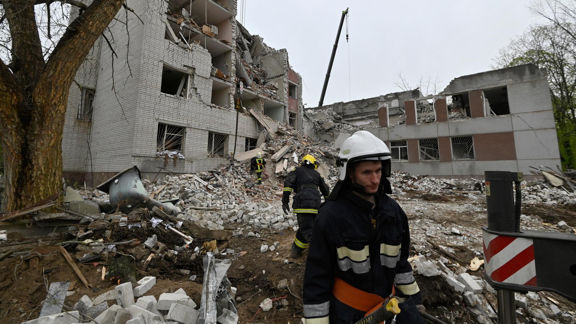 Zwei Feuerwehrleute arbeiten vor den Trümmern eines eingestürzten Hauses. Der Himmel ist bedeckt. 