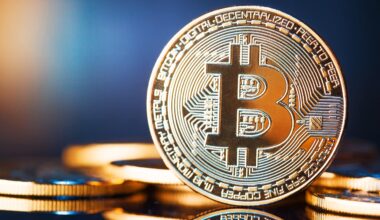 Nahaufnahme einer aufrecht stehenden physischen Bitcoin-Münze mit kreisförmigem Design, andere Münzen im Hintergrund, auf einer spiegelnden Oberfläche, die die besonderen Merkmale von Bitcoin hervorhebt.