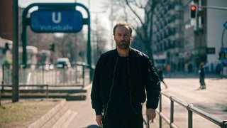 Kommissar Karow aus dem Tatort läuft auf einer Strasse durch Berlin, im Hintergrund eine U-Bahn-Station.