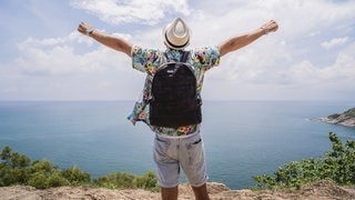 Mann steht an einer Küste und streckt die Arme raus, weil er sich freut, dass er alleine reist. 