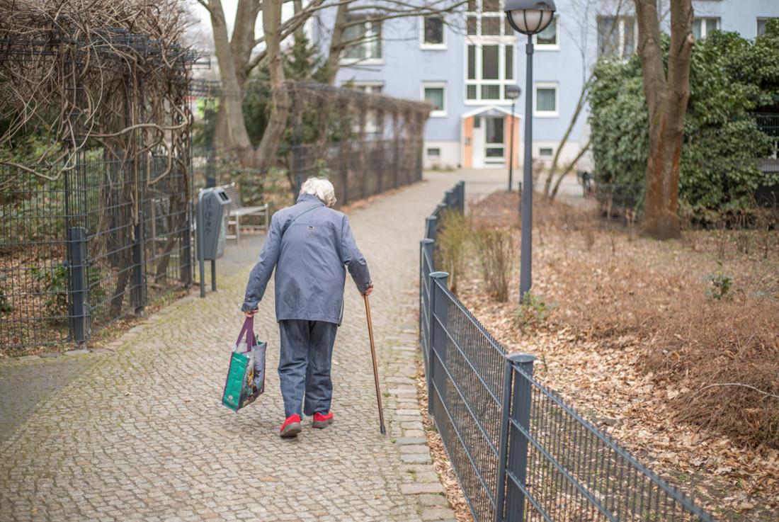 Eine ältere Frau mit Gehstock und Einkaufstasche läuft einen Weg entlang