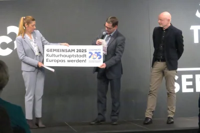 Chemnitz feiert Kulturhauptstadt-Abschluss: 2 Millionen Besuchende, Rekordzahlen und ein neues Selbstbewusstsein - Lößnitz’ Bürgermeister, Alexander Troll (m.), zog er ein großes Schild hinter der Bühne hervor, welches viele der Kommunen an ihre Ortseingangsschilder hingen und sich zur Kulturhauptstadtregion bekannten.