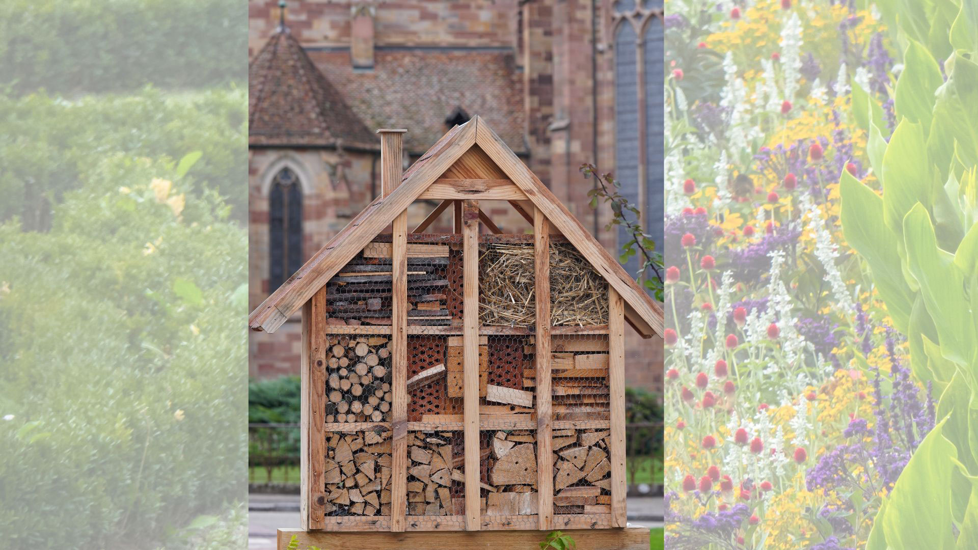 Ein großes Insektenhotel aus Holz steht im Freien. Es ist in mehrere Fächer unterteilt, die mit Naturmaterialien wie Holzstücken, Stroh und Ziegeln gefüllt sind. Im Hintergrund ist unscharf eine Kirchenfassade zu sehen.