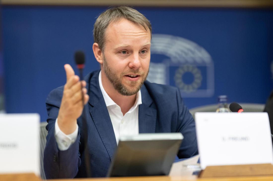 Daniel Freund sitzt an einem Tisch und spricht, während er seine rechte Hand hebt. 