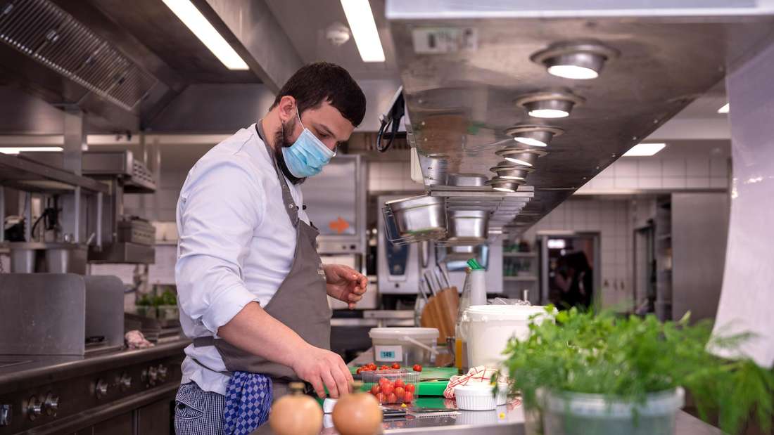 Ein Koch steht an einer Arbeitspfläche in einer Küche und richtet seine Zutaten, darunter sind Tomaten.