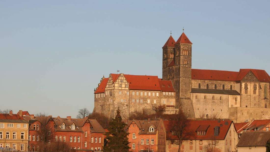 Eine Burg oder eine Kirche steht auf einer Anhöhe. Davor stehen Häuser. Die Sonne scheint unterzugehen. 