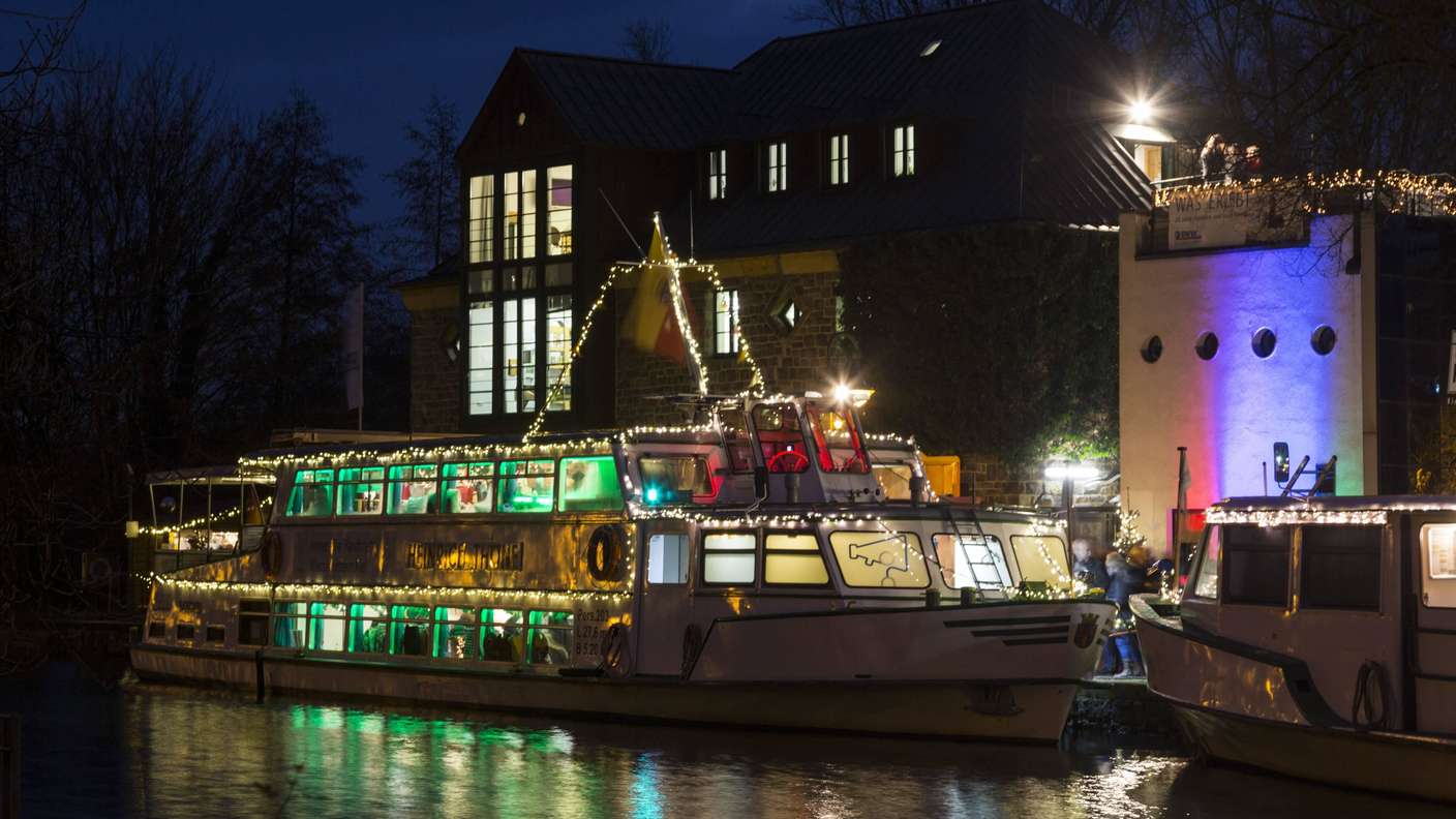 Ein festlich beleuchtetes Ausflugsschiff, das abends an einem Anleger vor einem modernen Wohnhaus mit großen Fenstern in Mülheim liegt, wobei sowohl das Boot als auch die umliegenden Gebäude weihnachtlich mit bunten Lichtern geschmückt sind.