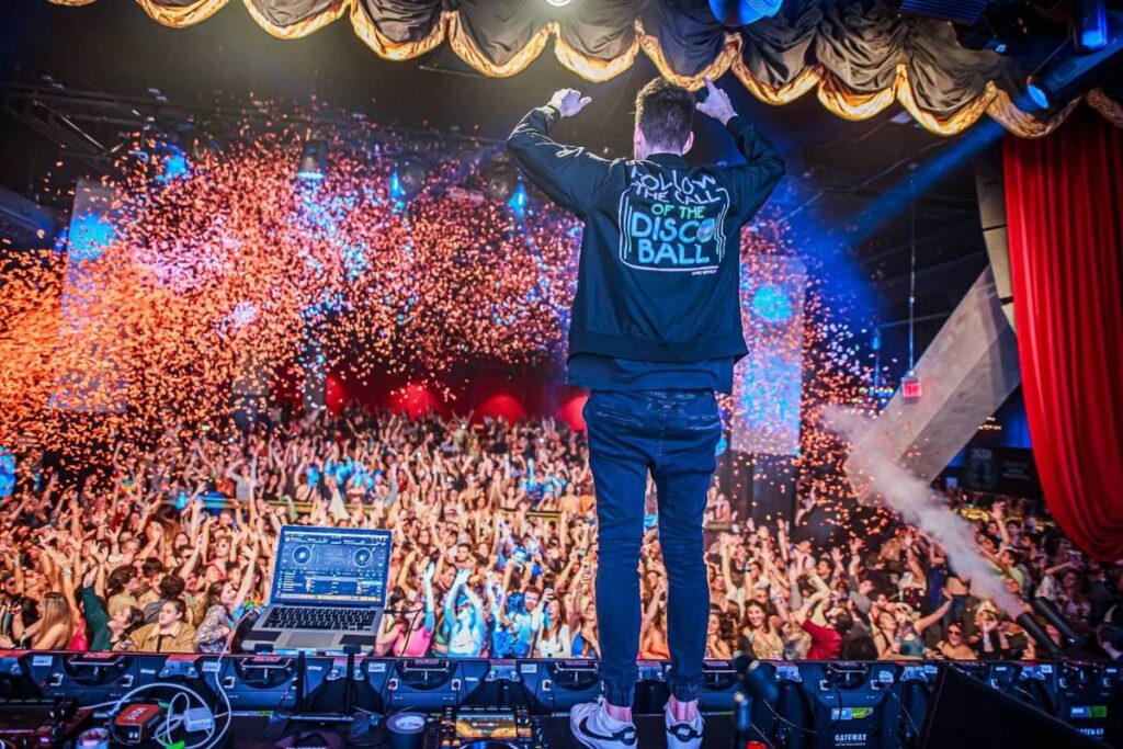 ein DJ steht mit erhobenen Armen auf der Bühne von Gimme Gimme Disco, Konfetti wird ins Publikum geschossen und die Leute auf der Tanzfläche jubeln mit ausgestreckten Armen
