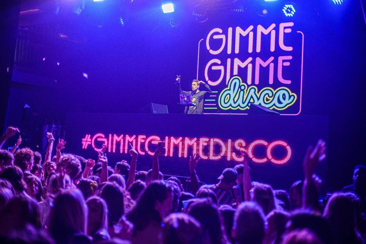 Leute tanzen auf der Tanzfläche von Gimme Gimme Disco während der DJ auf der Bühne hinter seinem Mischpult Musik spielt
