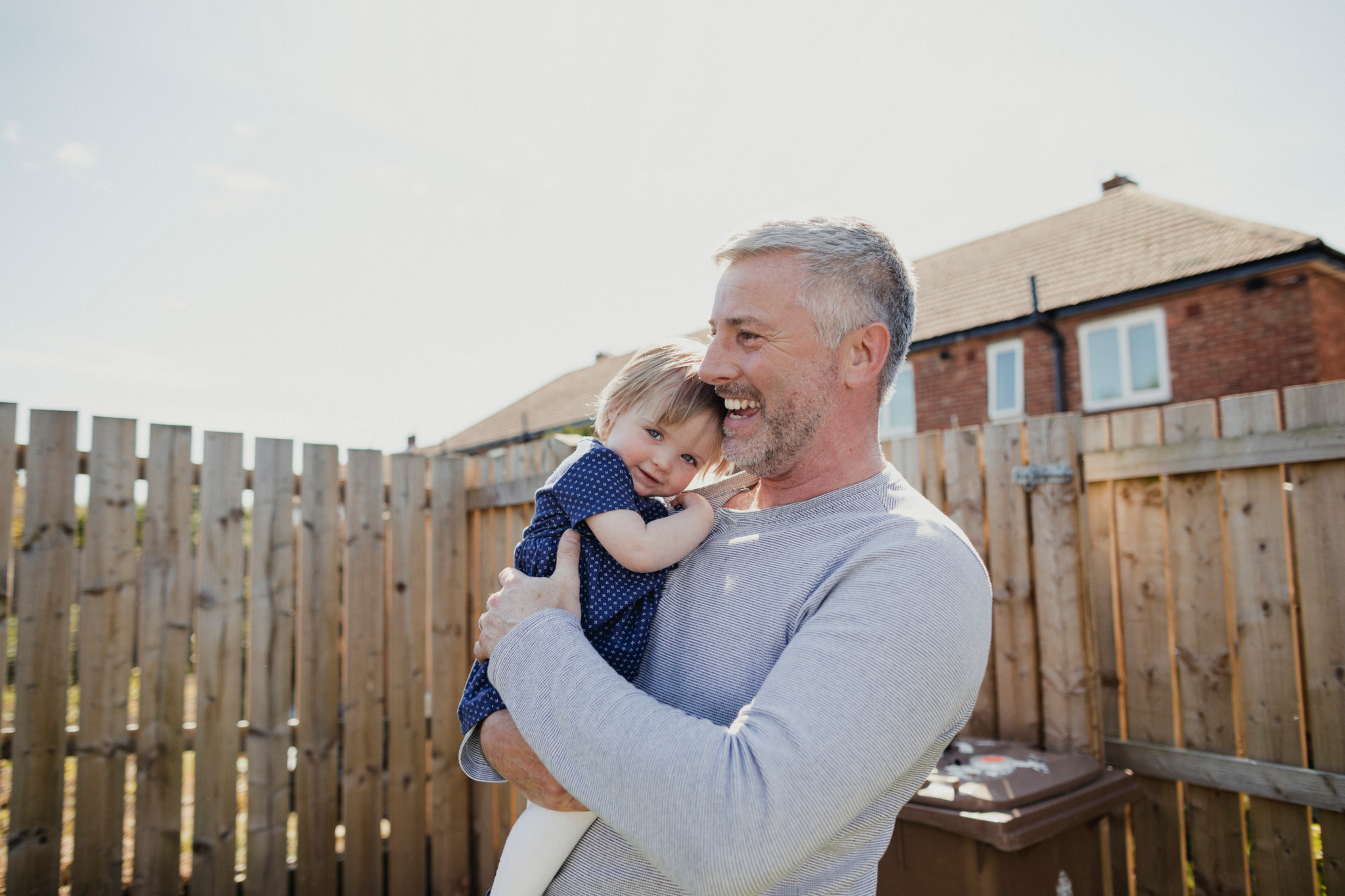 Reifer Vater hält seine kleine Tochter im Sonnenschein, während sie die Kamera anlächelt.