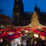 Stände mit roten Dächern und eine beleuchtete Fichte stehen auf dem Domplatz vor dem Dom.