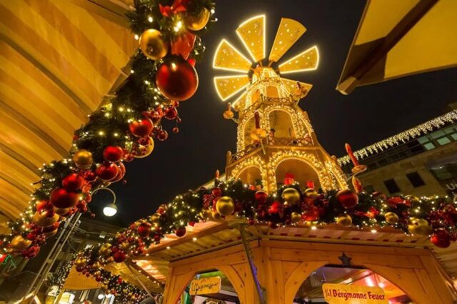 Eine festlich geschmückte Weihnachtsmarkt-Bude, umgeben von Lichtern und großen Ornamenten, mit einer beleuchteten Windmühle im Hintergrund.
