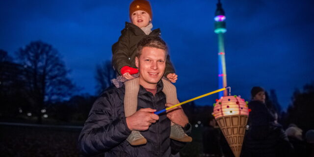 Ein Mann hat ein Kind auf den Schultern und hält eine Laterne zu St. Martin. Im Hintergrund ist der erleuchtete Florianturm im Dortmunder Westfalenpark zu sehen.
