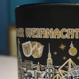 Die Glühwein-Tasse 2025 des Dortmunder Weihnachtsmarkts ist schwarz-gold und zeigt verschiedene Sehenswürdigkeiten der Stadt.