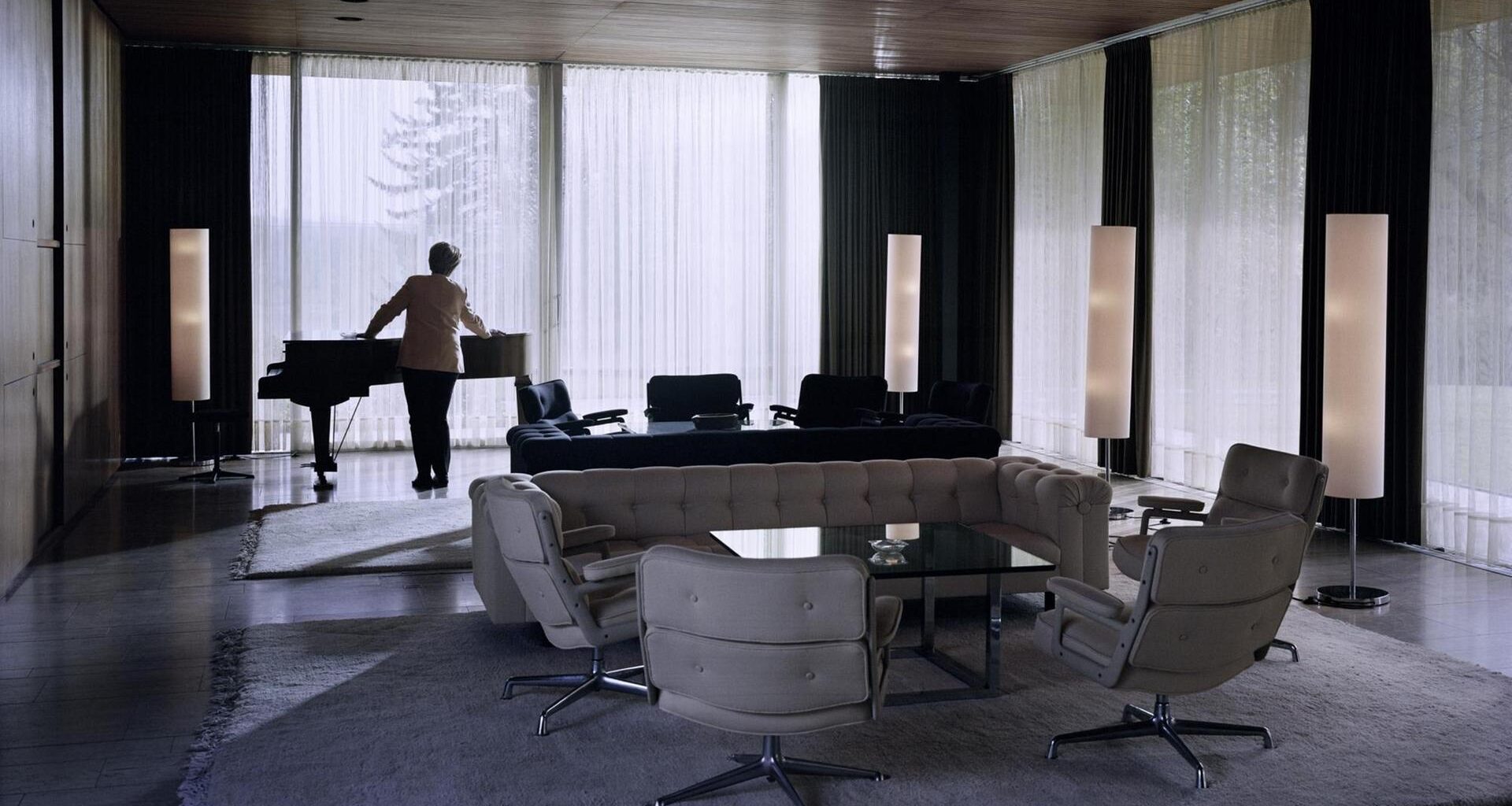 Eine Doppelgängerin von Angela Merkel steht an einem Flügel im Kanzlerbungalow.