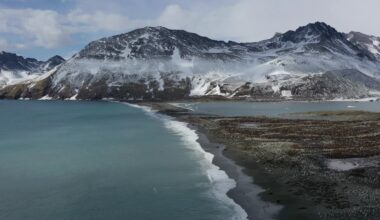 Eine Küstenlandschaft mit schneebedeckten Bergen im Hintergrund. Im Vordergrund erstreckt sich ein Strand entlang des türkisfarbenen Meeres. Die Küstenlinie verläuft diagonal durch das Bild, während Wolken den Himmel bedecken. Die Szene vermittelt eine ruhige, natürliche Atmosphäre.