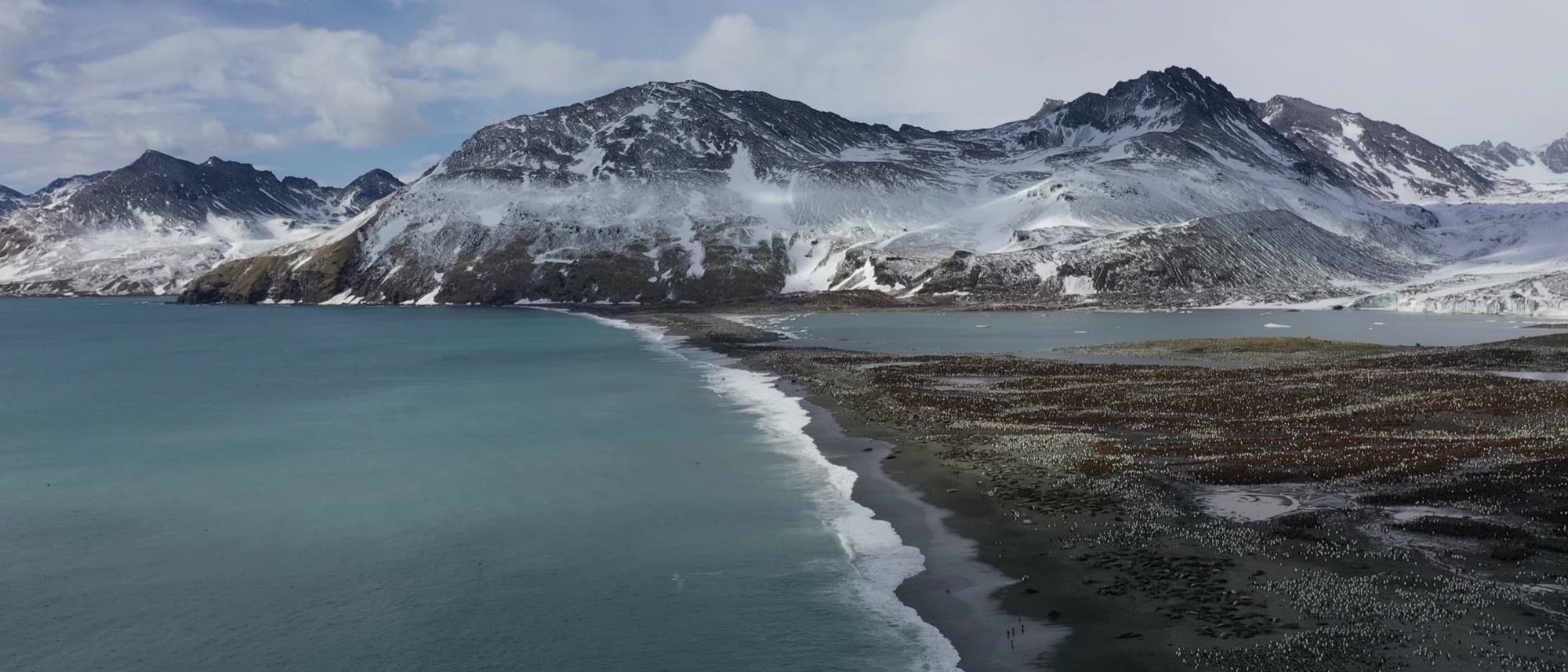 Eine Küstenlandschaft mit schneebedeckten Bergen im Hintergrund. Im Vordergrund erstreckt sich ein Strand entlang des türkisfarbenen Meeres. Die Küstenlinie verläuft diagonal durch das Bild, während Wolken den Himmel bedecken. Die Szene vermittelt eine ruhige, natürliche Atmosphäre. Eine Küstenlandschaft mit schneebedeckten Bergen im Hintergrund. Im Vordergrund erstreckt sich ein Strand entlang des türkisfarbenen Meeres. Die Küstenlinie verläuft diagonal durch das Bild, während Wolken den Himmel bedecken. Die Szene vermittelt eine ruhige, natürliche Atmosphäre.