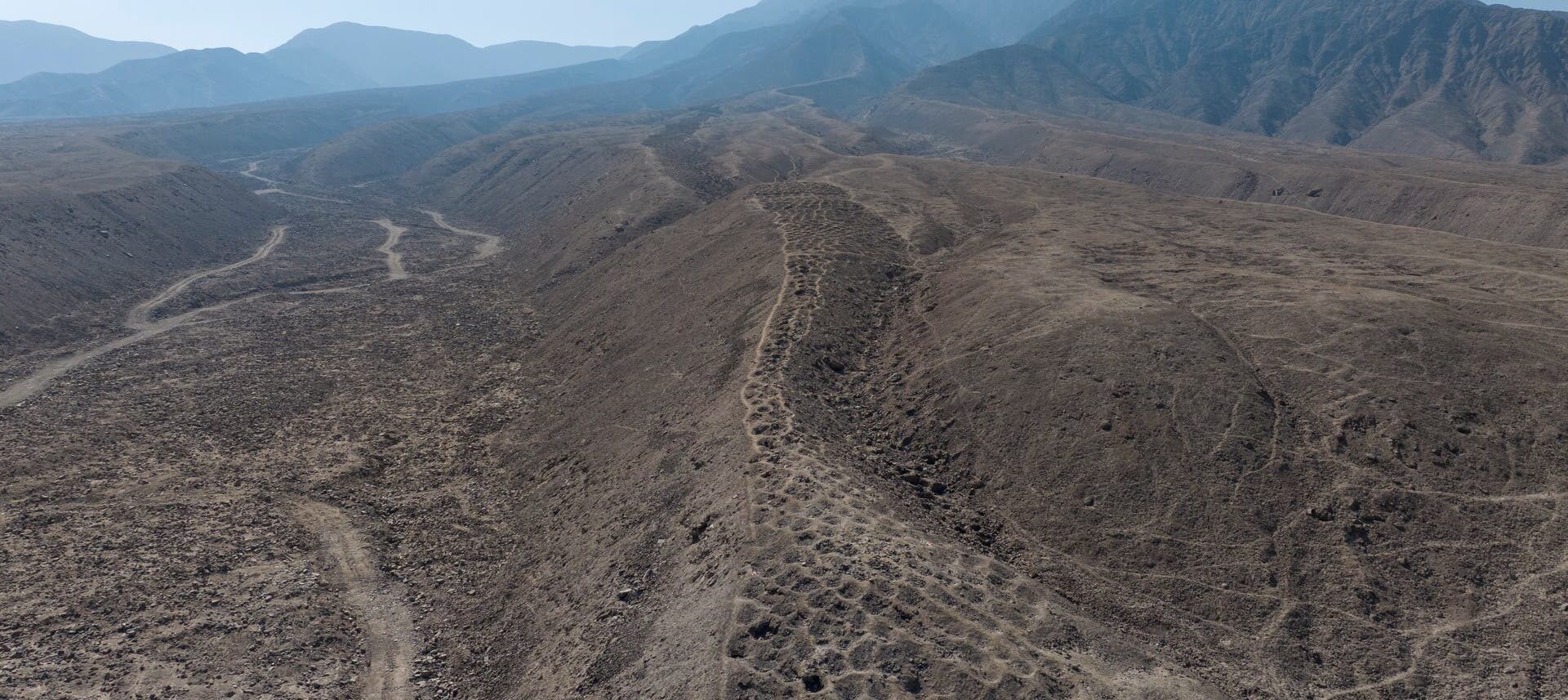 Luftaufnahme einer trockenen, hügeligen Landschaft mit erodierten Bergrücken und Tälern. Im Hintergrund sind grüne landwirtschaftliche Felder und ein Flussbett zu sehen, die die karge Wüstenlandschaft kontrastieren. Die Szene zeigt eine lange Reihe von künstlich angelegten Gruben.