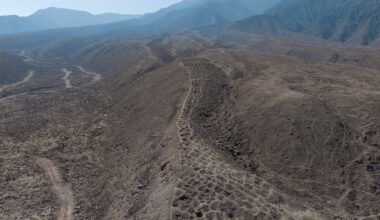 Luftaufnahme einer trockenen, hügeligen Landschaft mit erodierten Bergrücken und Tälern. Im Hintergrund sind grüne landwirtschaftliche Felder und ein Flussbett zu sehen, die die karge Wüstenlandschaft kontrastieren. Die Szene zeigt eine lange Reihe von künstlich angelegten Gruben.