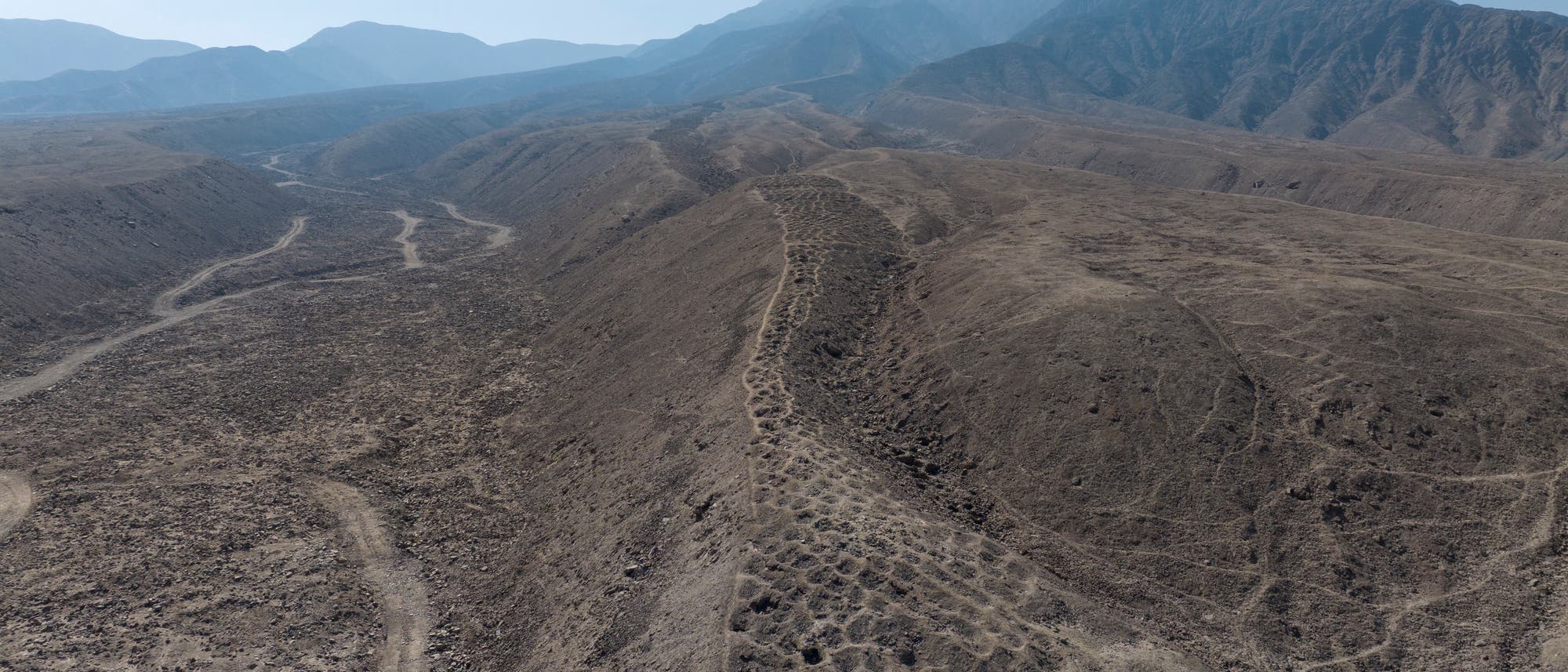Luftaufnahme einer trockenen, hügeligen Landschaft mit erodierten Bergrücken und Tälern. Im Hintergrund sind grüne landwirtschaftliche Felder und ein Flussbett zu sehen, die die karge Wüstenlandschaft kontrastieren. Die Szene zeigt eine lange Reihe von künstlich angelegten Gruben. Luftaufnahme einer trockenen, hügeligen Landschaft mit erodierten Bergrücken und Tälern. Im Hintergrund sind grüne landwirtschaftliche Felder und ein Flussbett zu sehen, die die karge Wüstenlandschaft kontrastieren. Die Szene zeigt eine lange Reihe von künstlich angelegten Gruben.