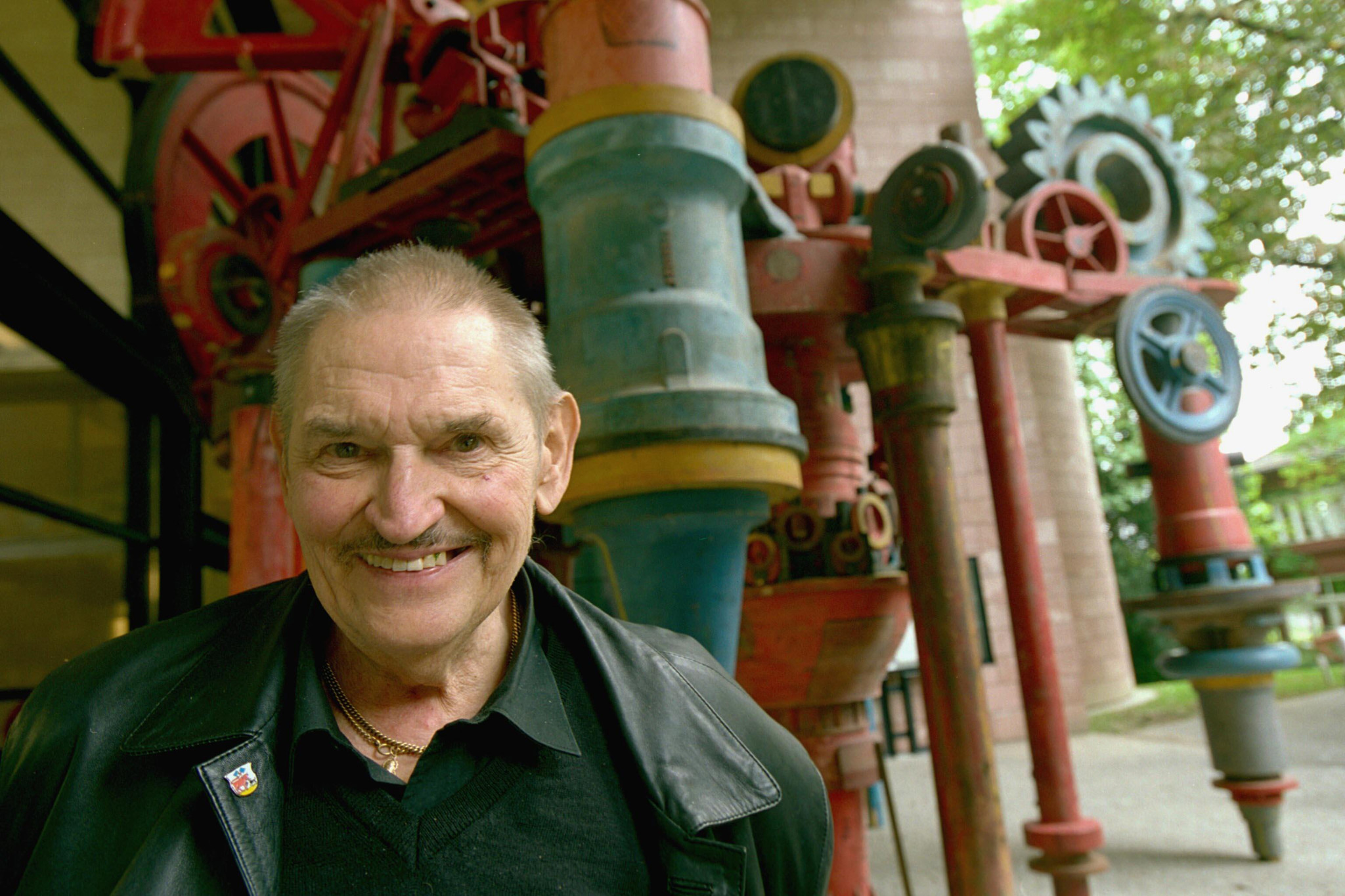 Bernhard Luginbühl vor einer seiner Skulpturen im Tinguély-Museum Basel, mechanische und bunte Elemente im Hintergrund.