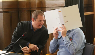 Angeklagter, der sich einen weißen Ordner vor das Gesicht hält, spricht mit seinem Anwalt im Gerichtssaal.