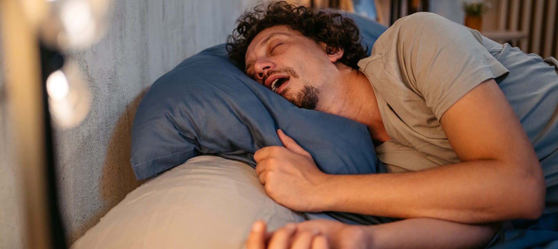 Eine Person liegt schlafend auf der Seite in einem Bett mit offenem Mund. Im Hintergrund ist eine sanfte Beleuchtung zu sehen.