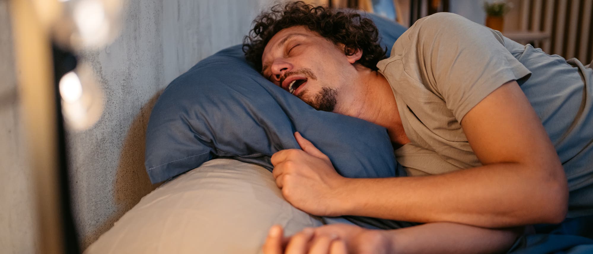 Eine Person liegt schlafend auf der Seite in einem Bett mit offenem Mund. Im Hintergrund ist eine sanfte Beleuchtung zu sehen. Eine Person liegt schlafend auf der Seite in einem Bett mit offenem Mund. Im Hintergrund ist eine sanfte Beleuchtung zu sehen.