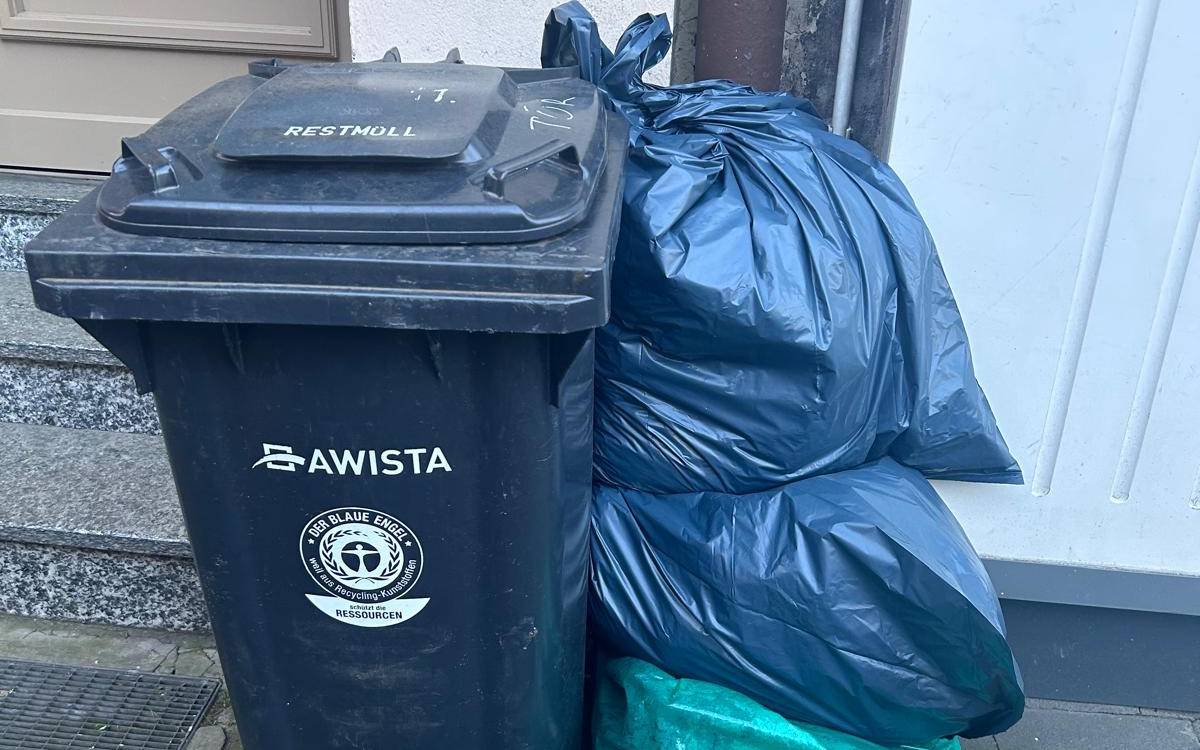 Eine Restmülltonne steht vor einer Hauswand, daneben mehrere verschlossene Säcke mit Restmüll.